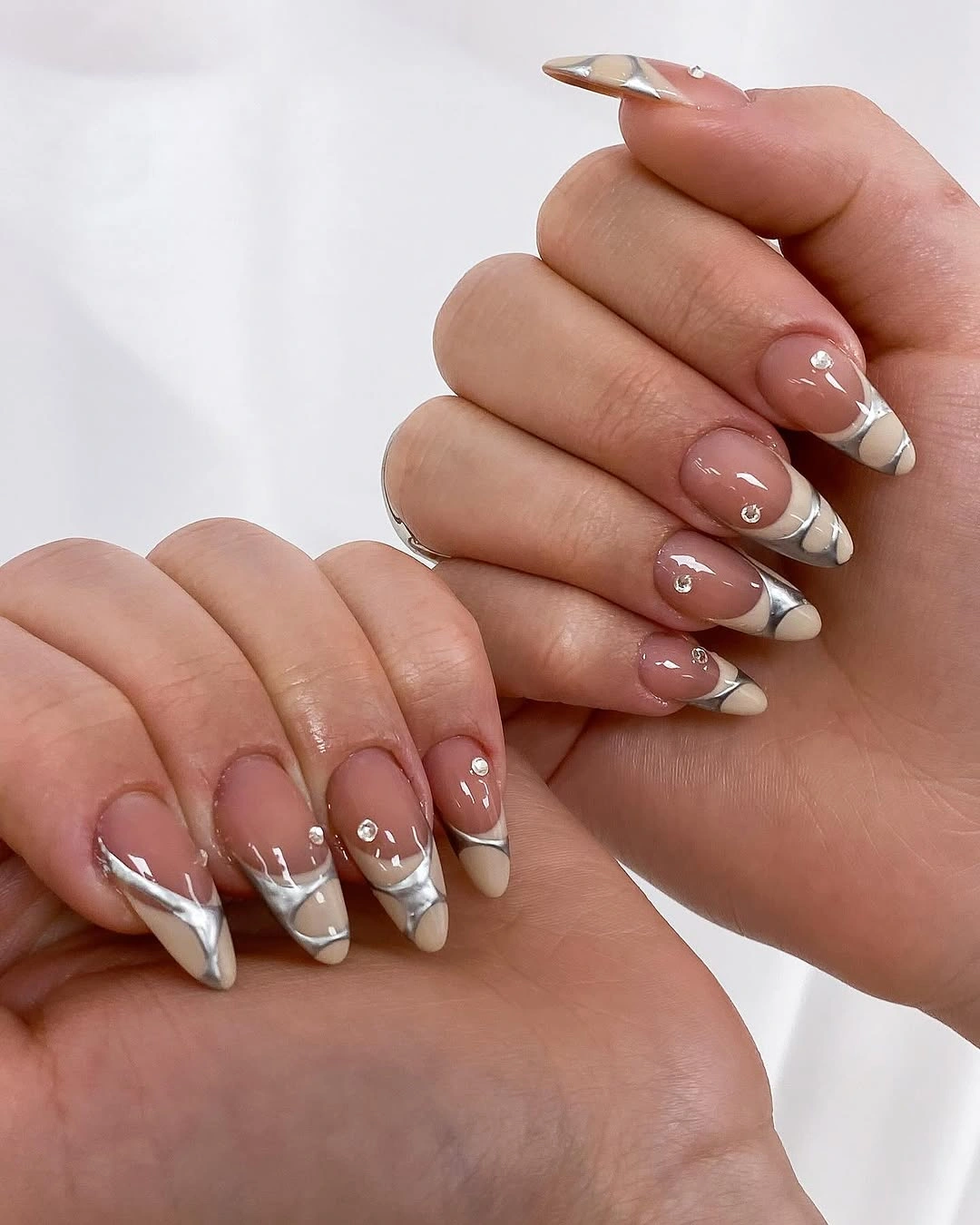 Pointed almond nails with nude bases, angled white tips, metallic-looking curves, and reflective accents